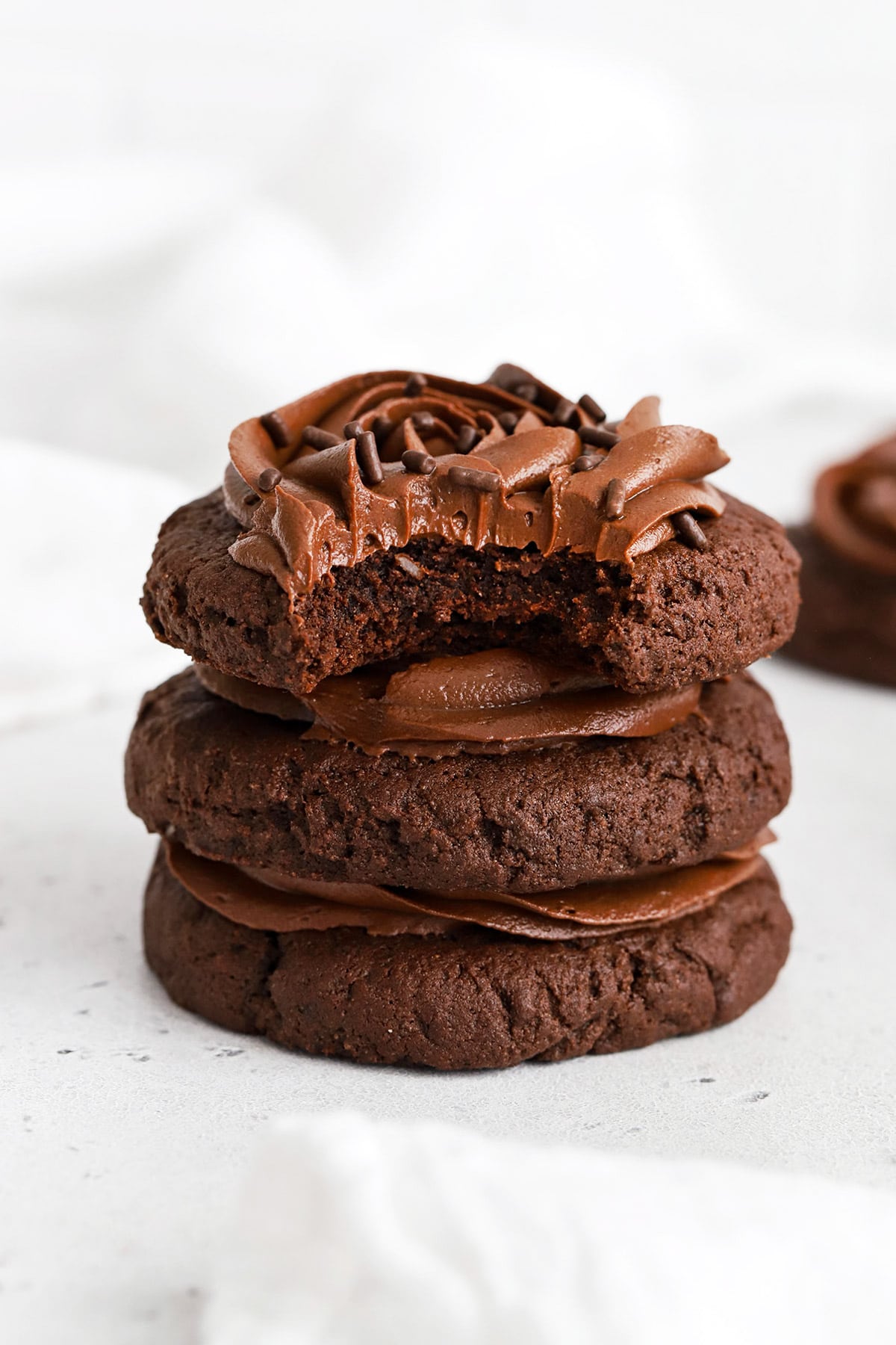 Front view of a stack of gluten-free crumbl chocolate cake cookies with chocolate frosting. One cookie has a bite taken out of it revealing the soft cakey center.