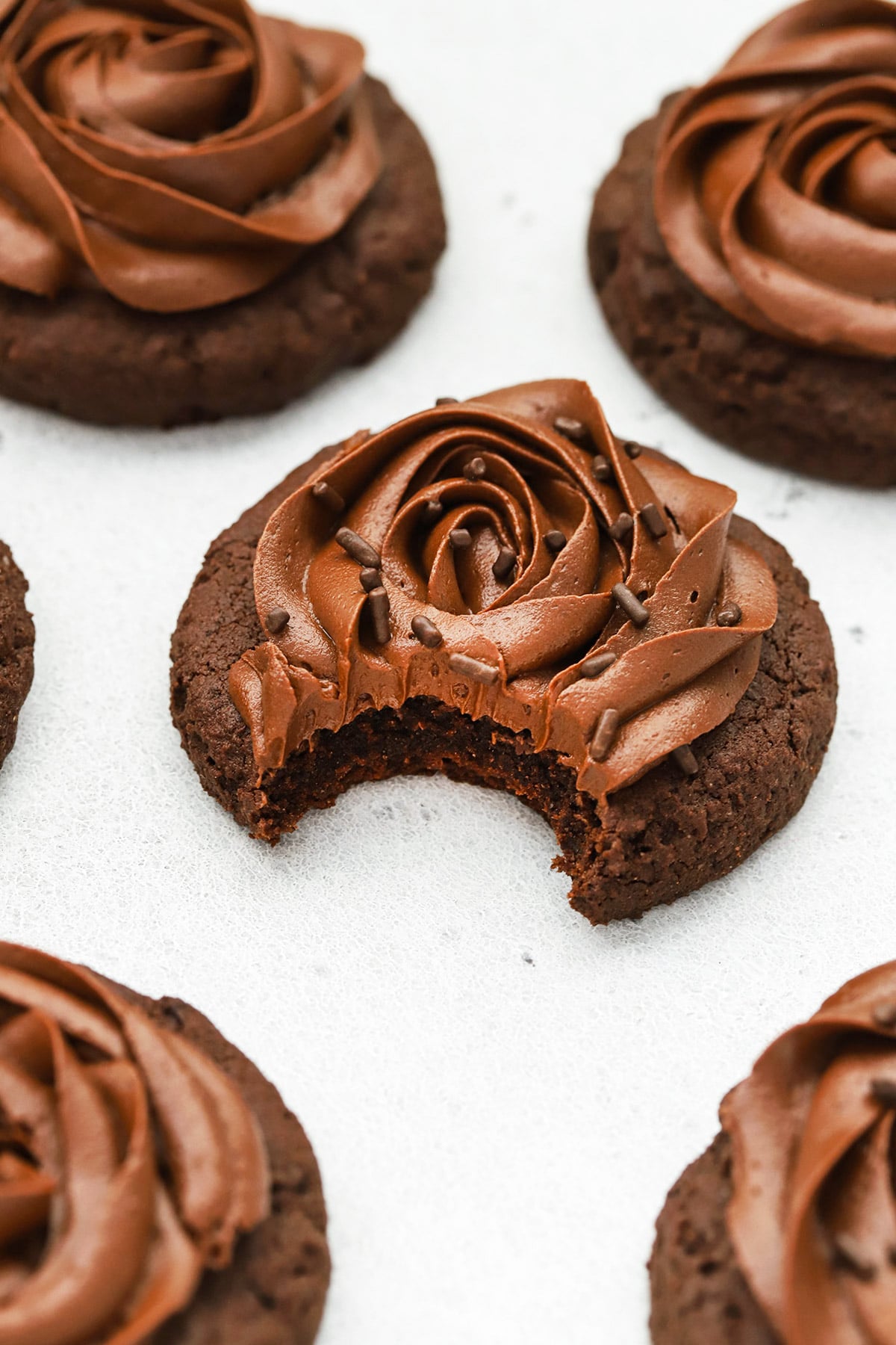 Front view of gluten-free crumbl chocolate cake cookies with chocolate frosting. One cookie has a bite taken out of it revealing the soft cakey center.