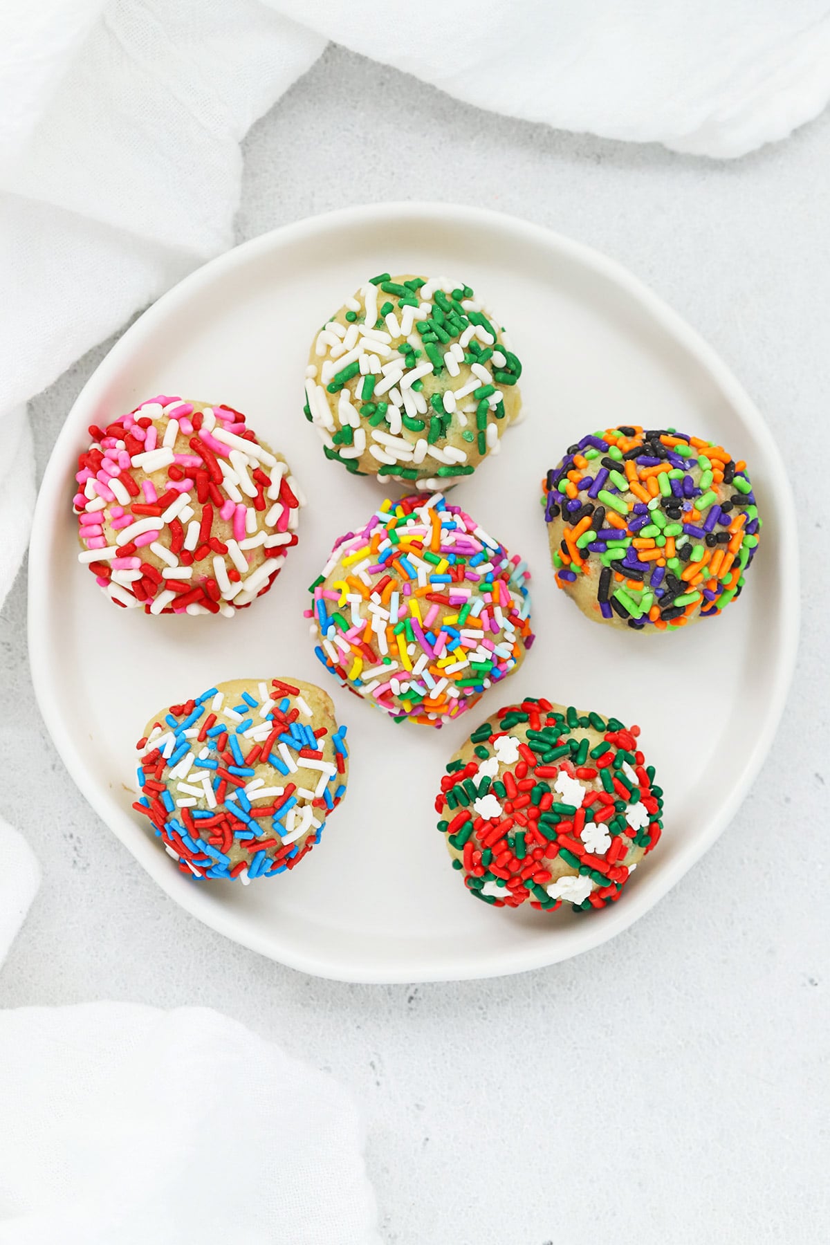 Overhead view of six gluten-free sprinkle sugar cookie dough balls rolled in different colored sprinkles for each holiday--pink, red and white for valentine's day, white and green for st. patrick's day, pastels for Easter, red white and blue for 4th of July, green, black, orange, and purple for halloween, and red, white and green for christmas.