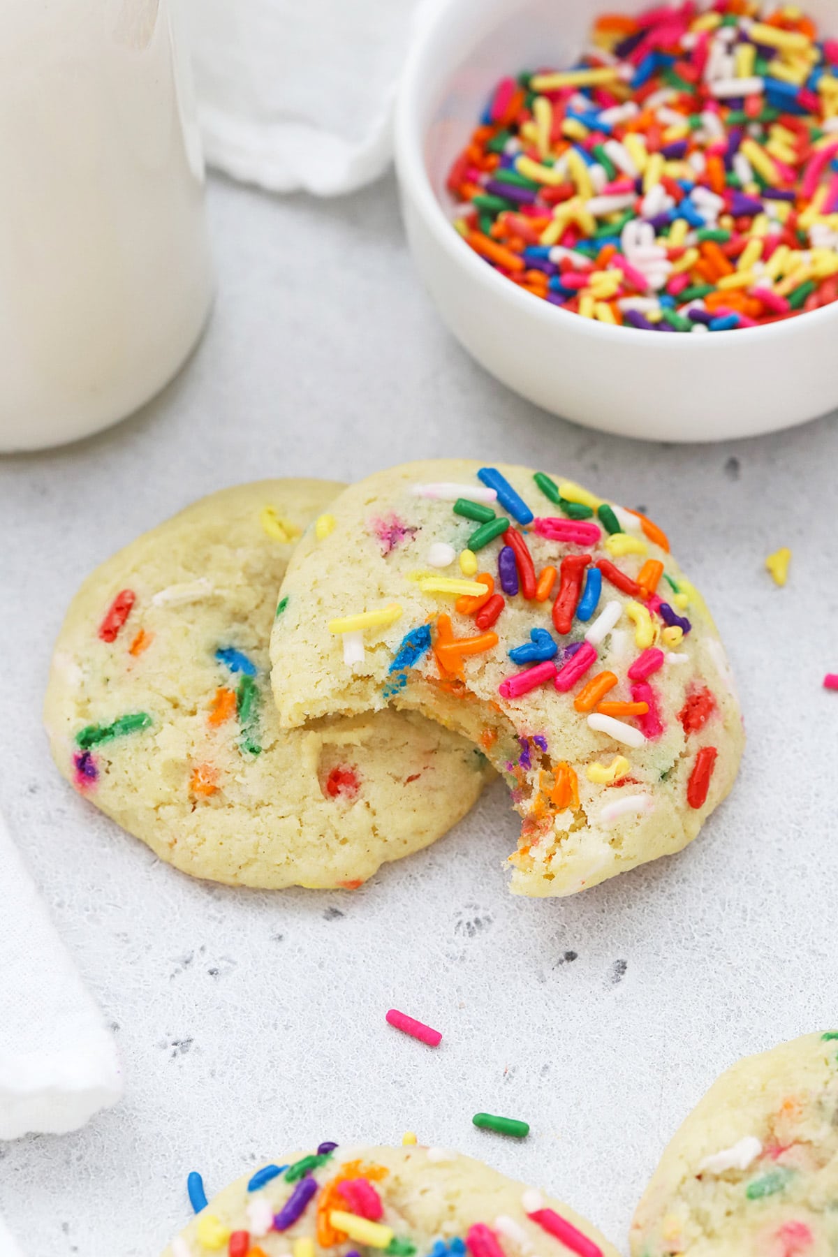 Gluten-free sprinkle sugar cookies stacked on top of each other. One has a bite taken out, revealing a soft gluten-free sugar cookie center