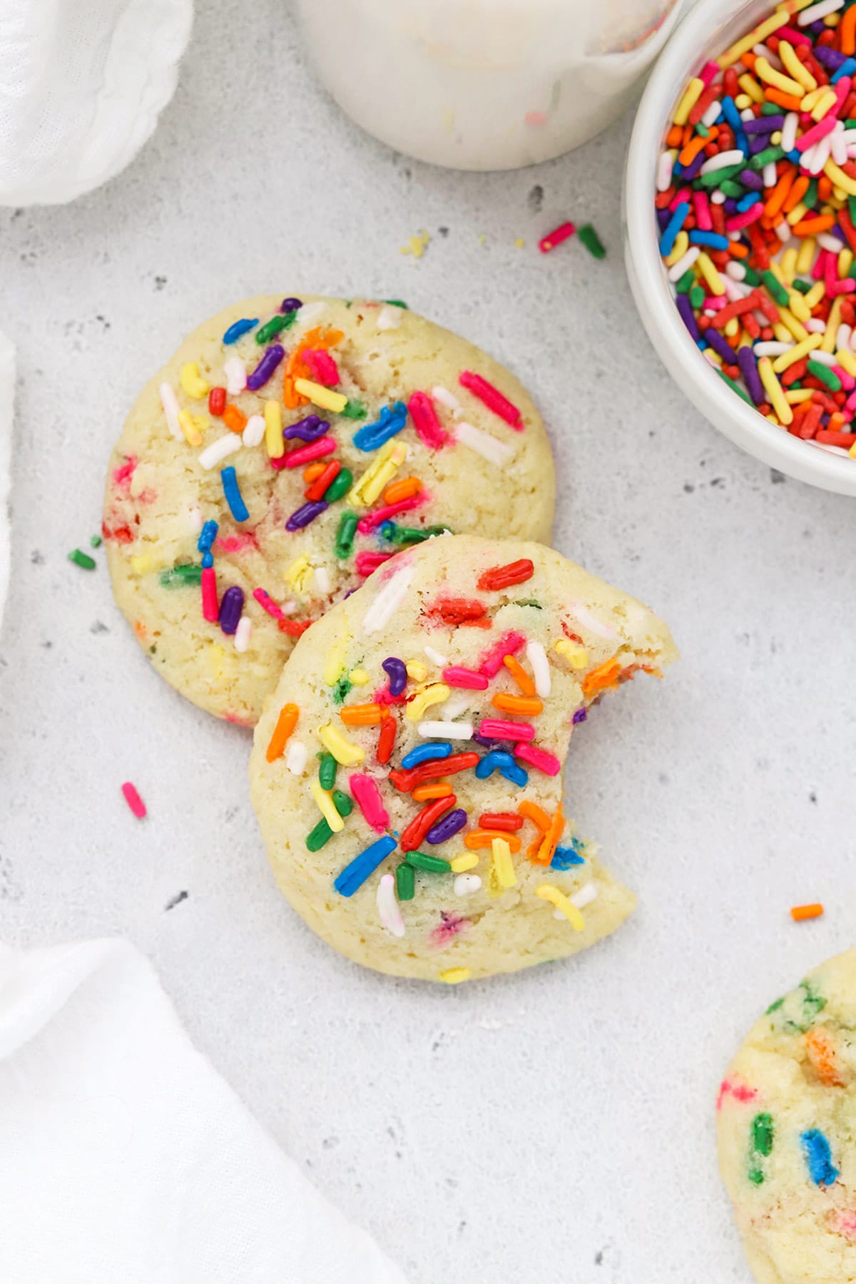 Gluten-free sprinkle sugar cookies stacked on top of each other. One has a bite taken out, revealing a soft gluten-free sugar cookie center