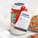 Front view of gluten-free holiday cookies in a jar with free printable gift tag with baked gluten-free m&m cookies in the background