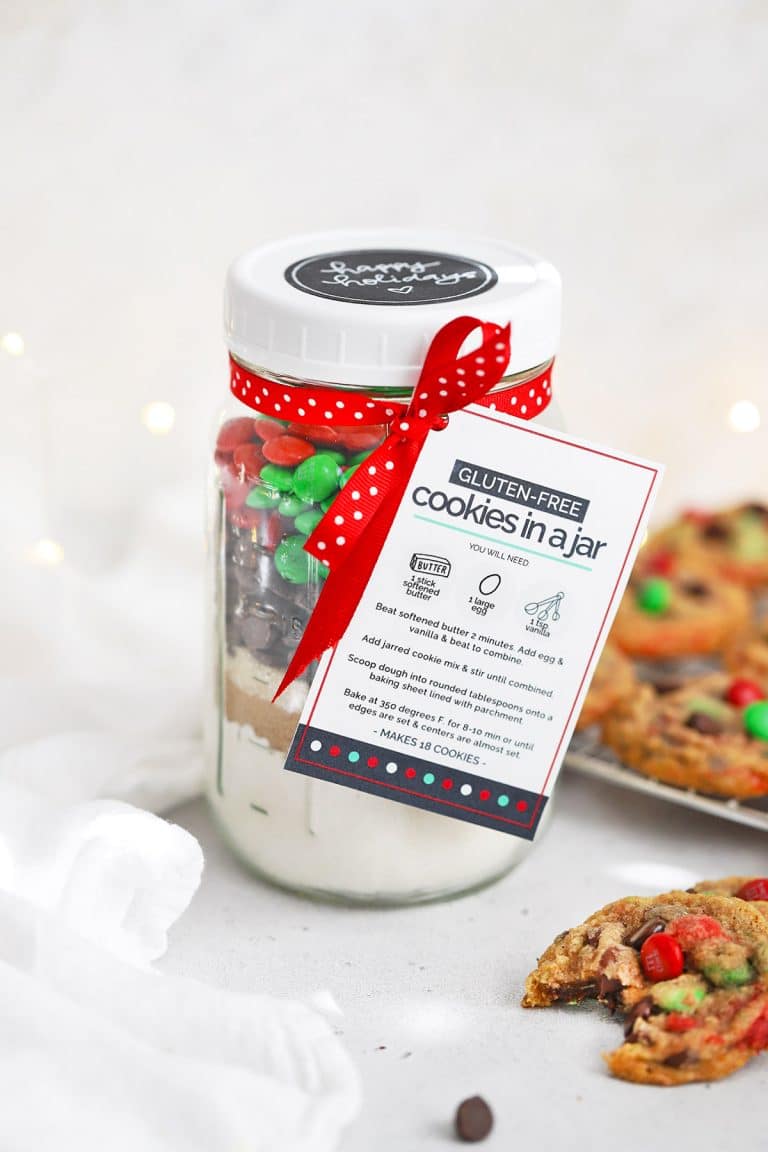 Front view of gluten-free holiday cookies in a jar with free printable gift tag with baked gluten-free m&m cookies in the background