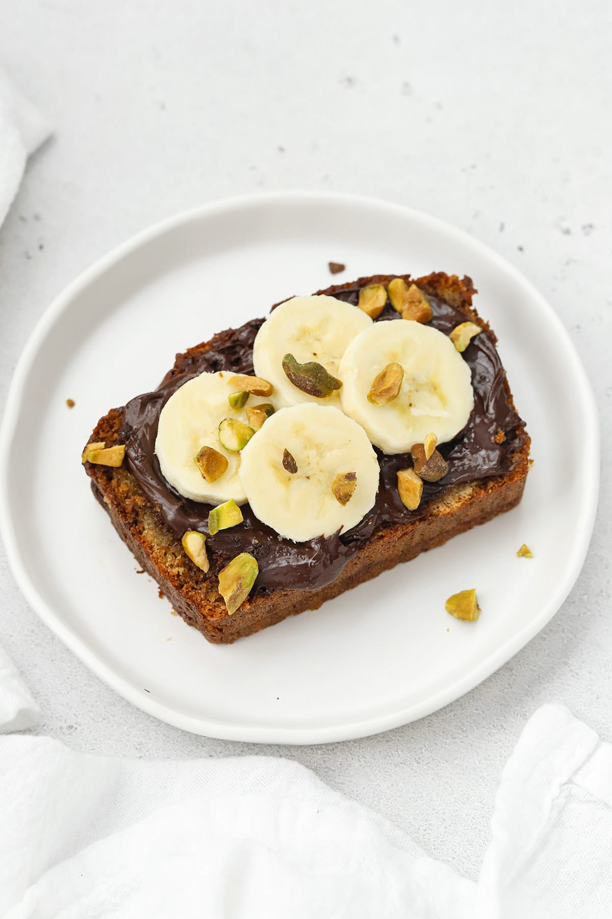 Slice of gluten-free banana bread topped with nutella, sliced bananas, and pistachios
