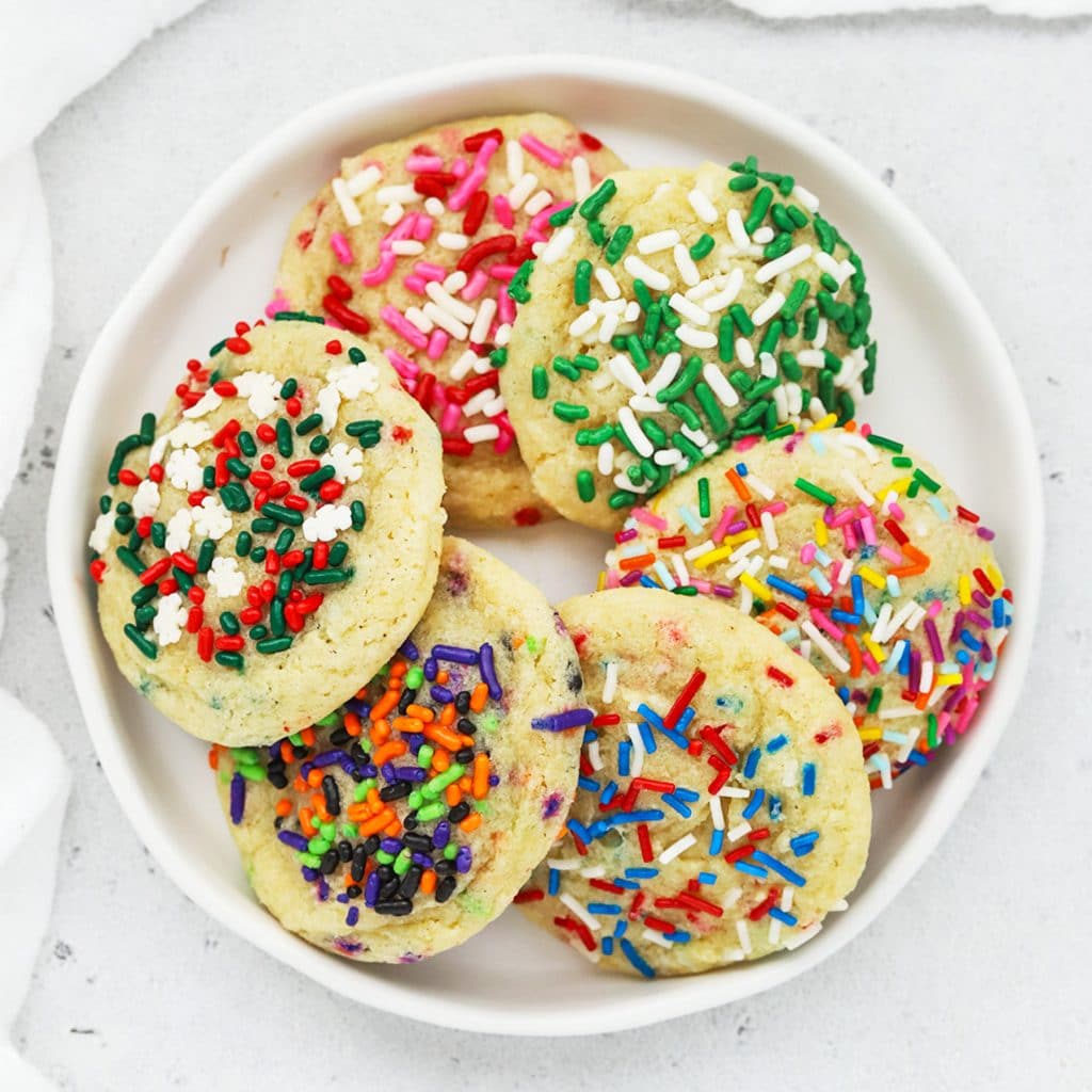 Overhead view of six gluten-free sprinkle sugar cookies with different colored sprinkles for each holiday--pink, red and white for valentine's day, white and green for st. patrick's day, pastels for Easter, red white and blue for 4th of July, green, black, orange, and purple for halloween, and red, white and green for christmas.