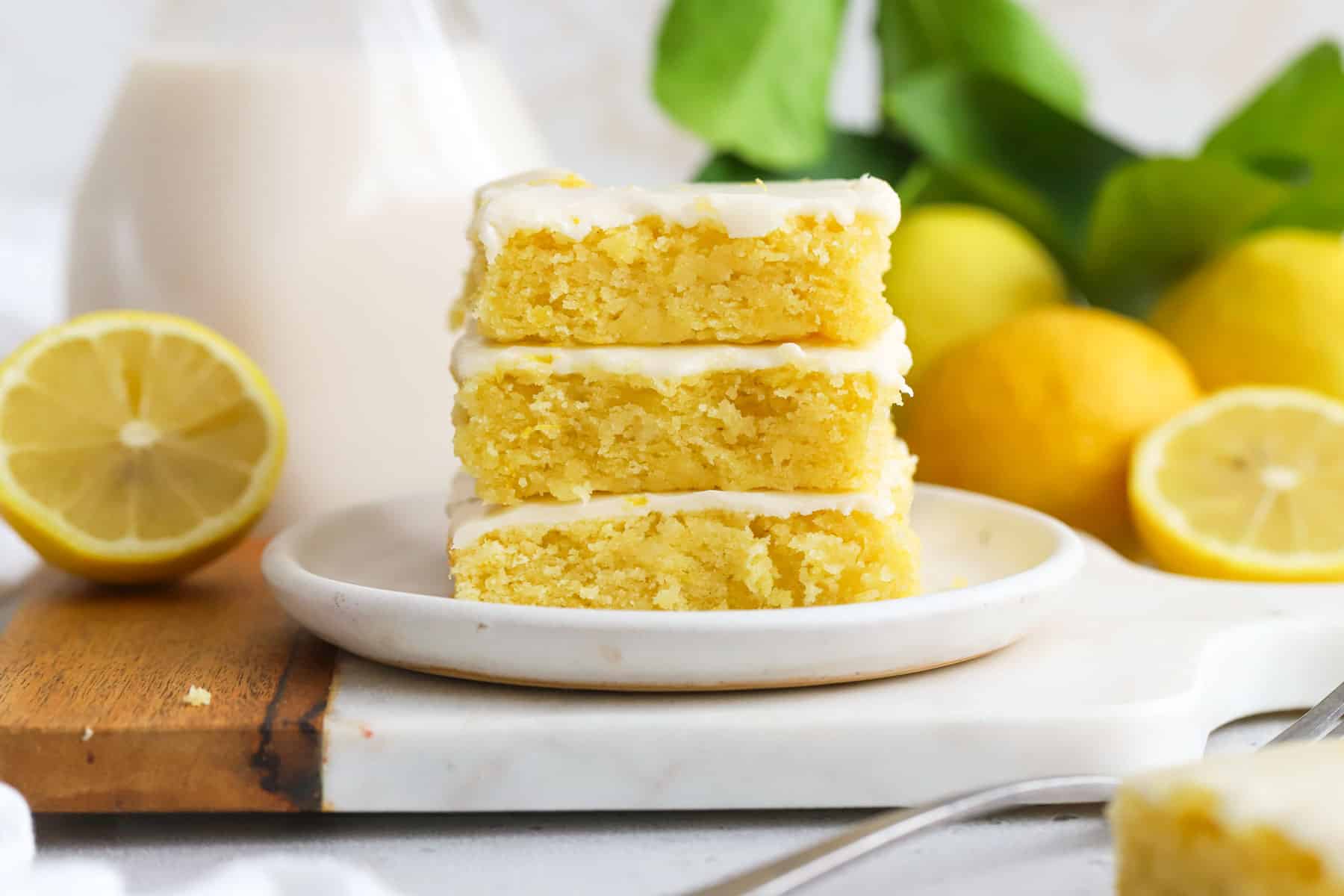 Three gluten-free lemon brownies stacked on a white dessert plate next to a bottle of milk