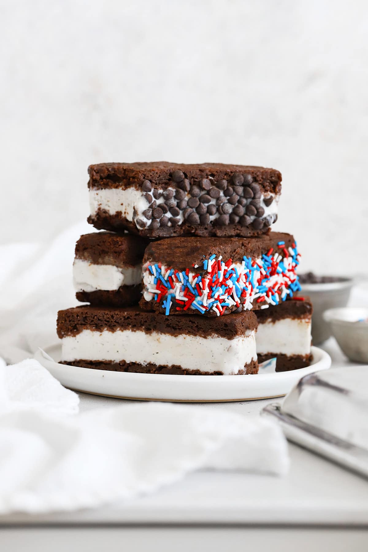 Gluten free ice cream sandwiches stacked on a plate. One is rolled in mini chocolate chips and one is rolled in red, white, and blue jimmies sprinkles