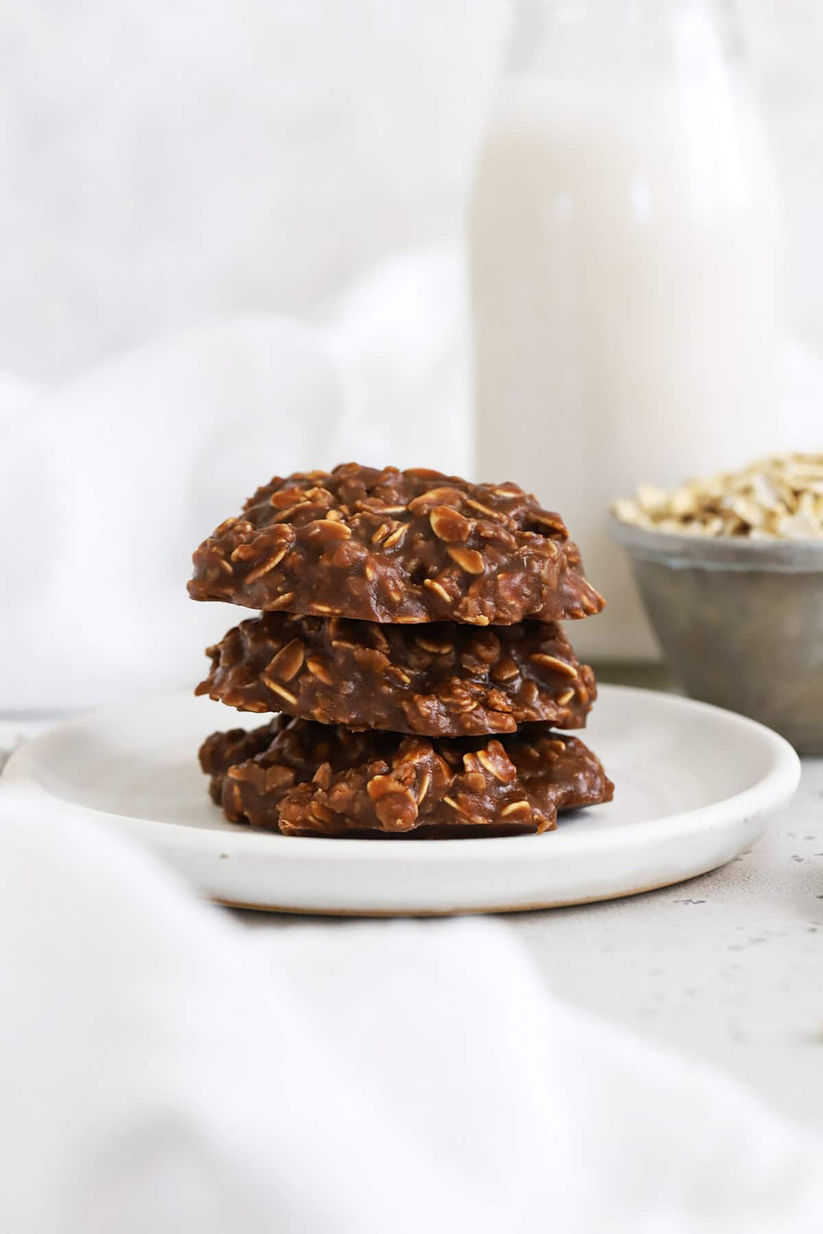 A stack of three gluten-free no-bake cookies on a white plate with a bottle of milk in the background