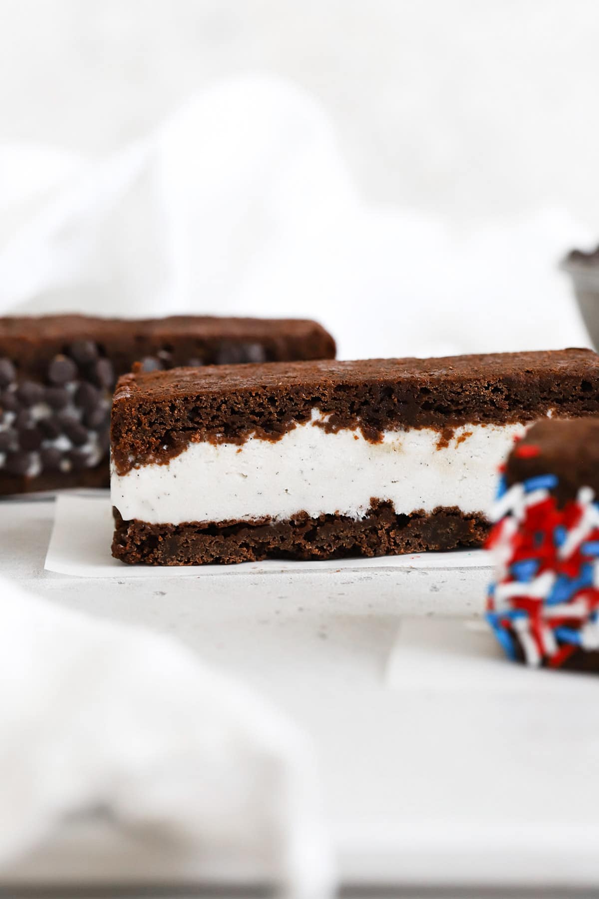 Three gluten-free brownie ice cream sandwiches on white background. One is rolled in chocolate chip and one is rolled in red, white, and blue sprinkles