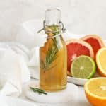 A small glass bottle of rosemary simple syrup with a plate of colorful citrus in the background