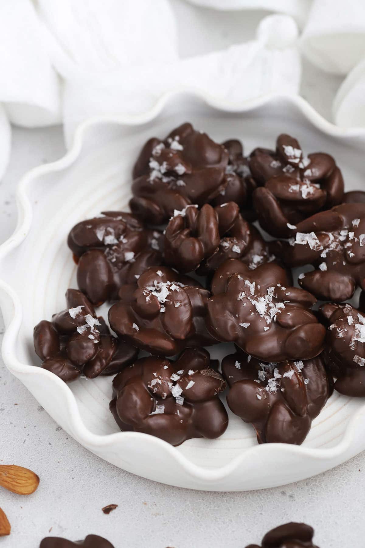 Front view of a ruffled white plate of dark chocolate almond clusters topped with flaky sea salt