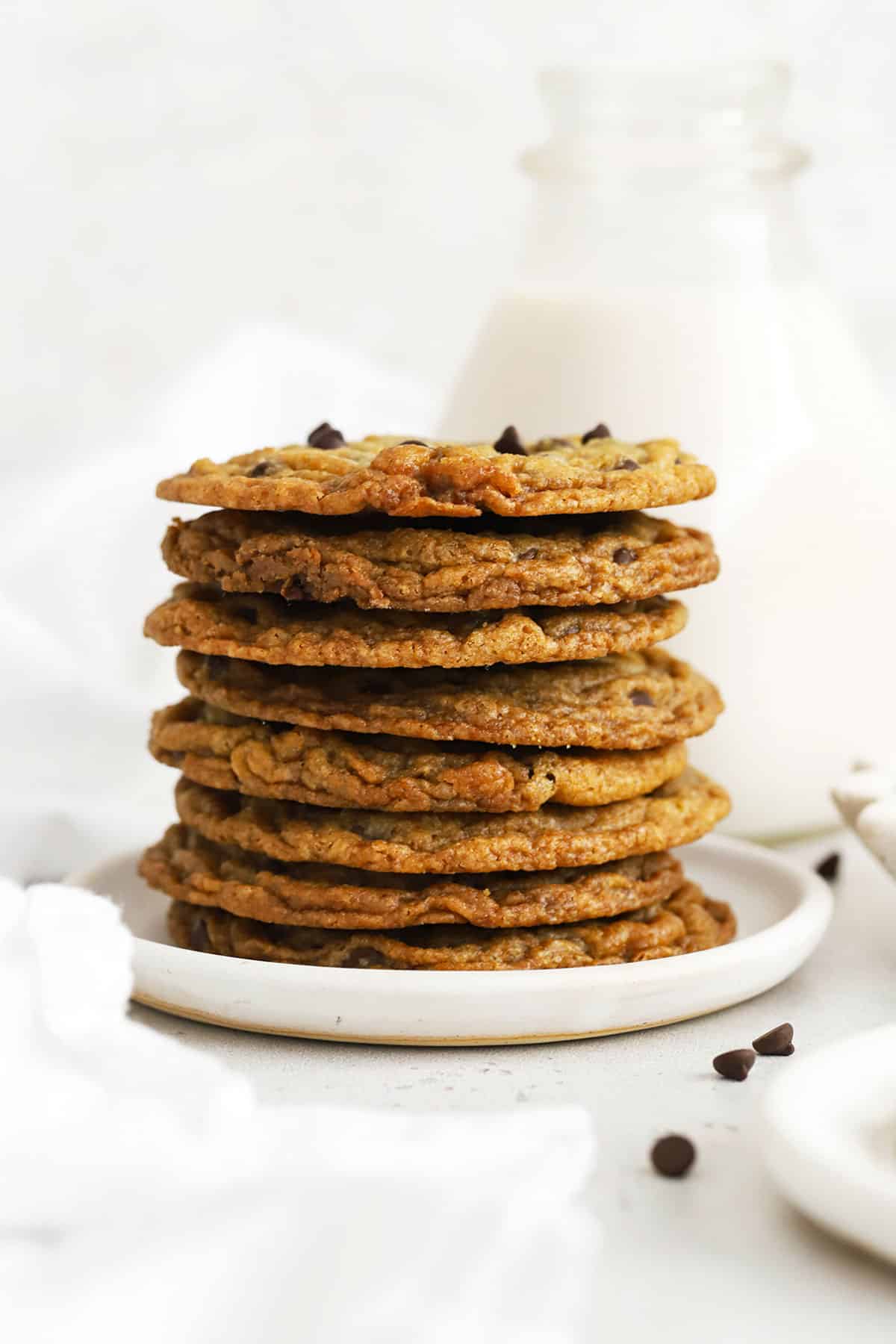 a stack of thin, crispy gluten-free chocolate chip cookies with a jar of milk in the background