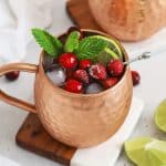 two copper mugs with cranberry moscow mule mocktails on wood and marble coasters