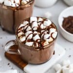 two glass mugs of gluten-free hot chocolate with marshmallows, chocolate syrup, and chocolate shavings
