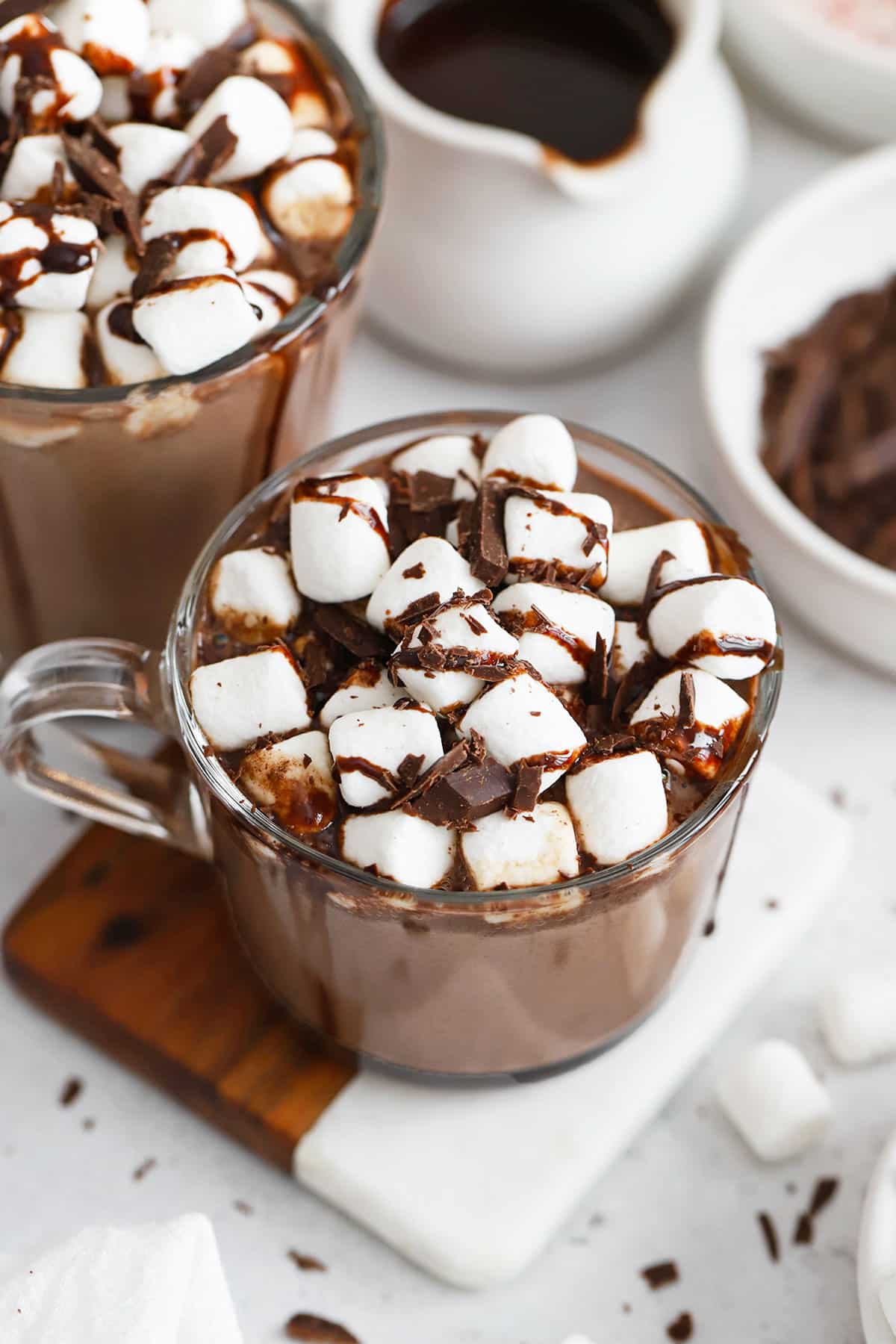 two glass mugs of gluten-free hot chocolate with marshmallows, chocolate syrup, and chocolate shavings