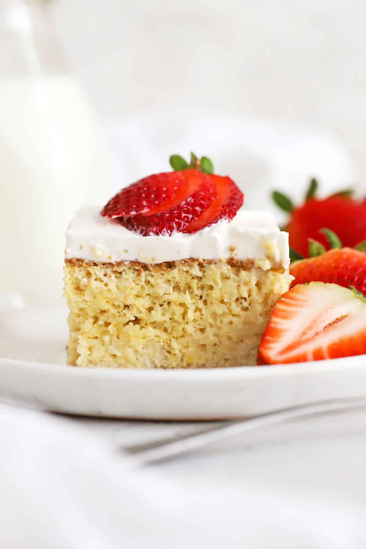 A slice of gluten-free tres leches cake topped with fresh strawberries on a white plate