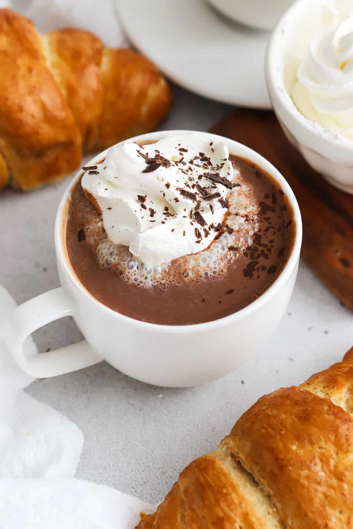 A cup of French hot chocolate topped with whipped cream, next to gluten-free croissants