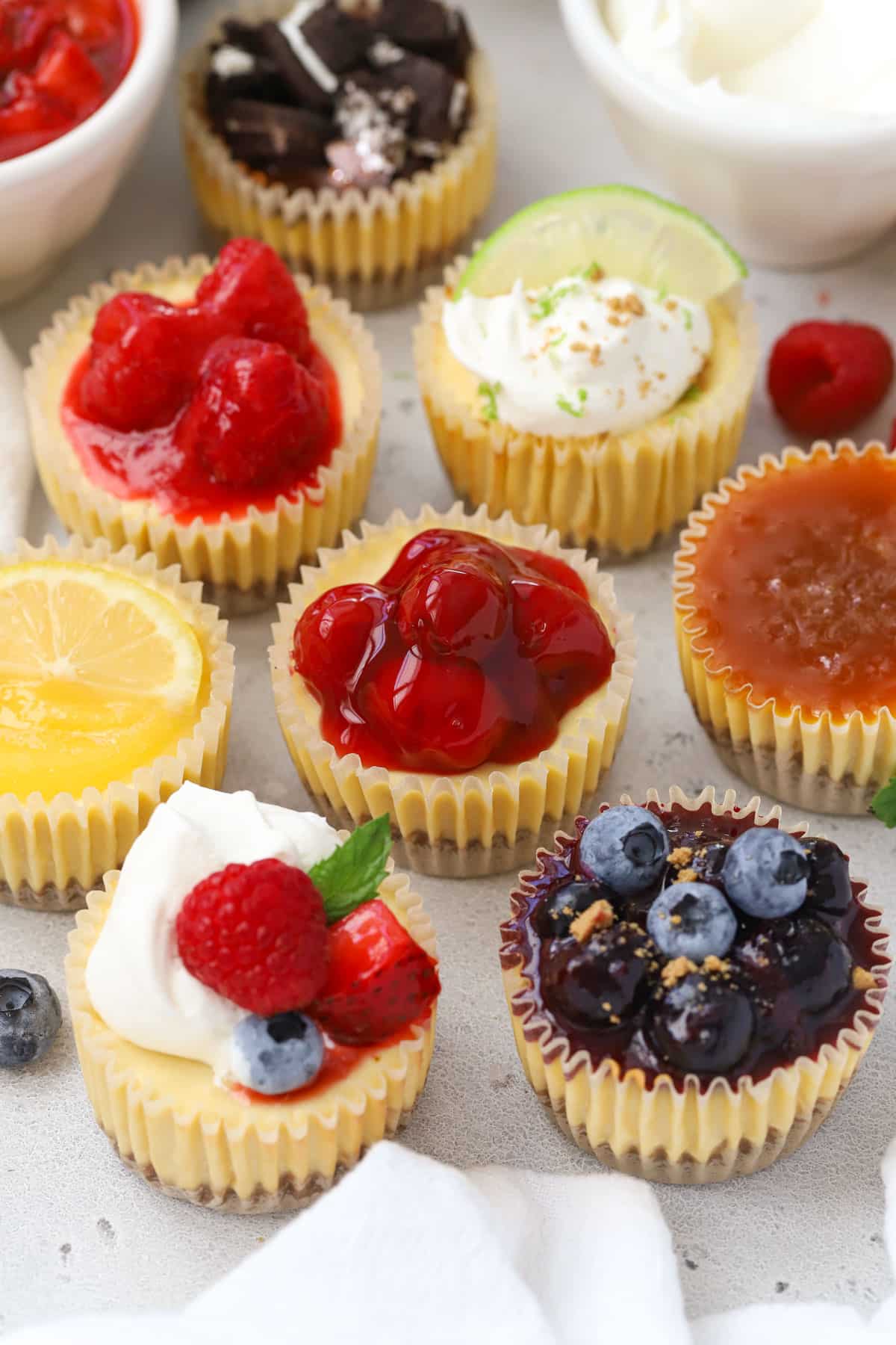 9 Flavors of mini gluten-free cheesecakes with different toppings on a white background